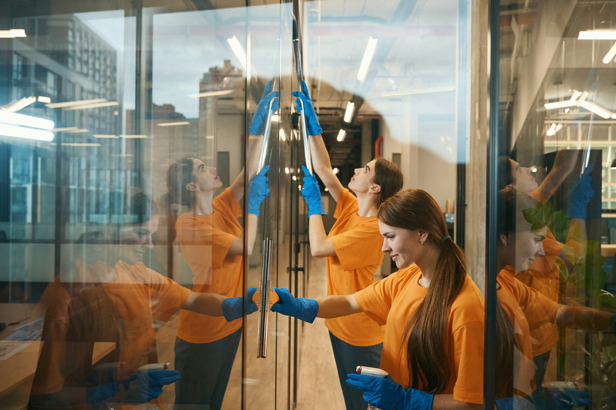 Young cleaners clean mirrored windows coworking spacer, they in simple uniforms